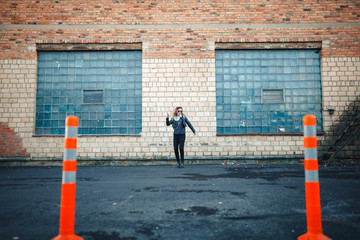 Young beautiful sexy woman with headphones and smartphone in hand in mirrored sunglasses, a black leather jacket, black jeans dancing by the wall of the industrial building of brick on the street