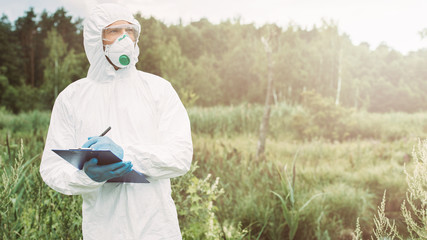 male scientist in protective mask, googles and suit looking away and writing in clipboard in meadow