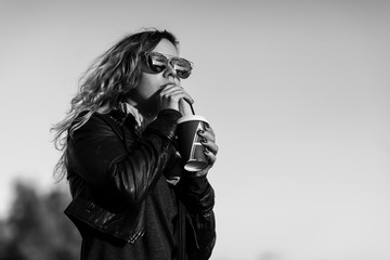 Attractive woman in mirrored sunglasses, a black leather jacket, drinking coffee on the waterfront river in the city in front the setting sun. Waving hair. city,bridge. Black and white picture.