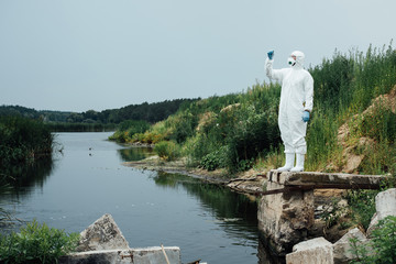Fototapeta premium male scientist in protective mask and suit looking at sample of water in test flask outdoors