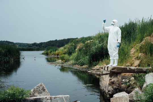 Male Scientist In Protective Mask And Suit Looking At Sample Of Water In Test Flask Outdoors