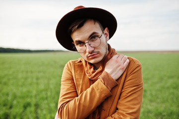 Stylish man in glasses, brown jacket and hat posed on green field.