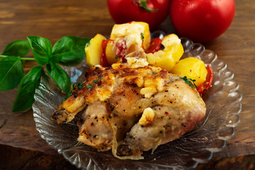 A simple meat dish of baked chicken with vegetables, cheese and green Basil on a round glass plate, next to two large red tomatoes.