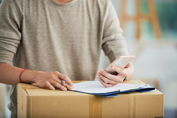 Crop view of man in casual clothes standing with pen at delivered packed cardboard box with clipboard and working with mobile phone in hand on blurred background .