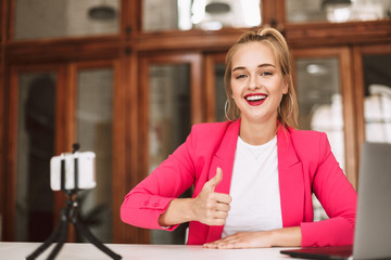 Beautiful smiling girl in pink jacket happily looking in camera showing thumb up while recording...