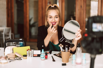 Beautiful smiling beauty blogger happily looking in camera holding lipstick and mirror in hands while recording new video for vlog
