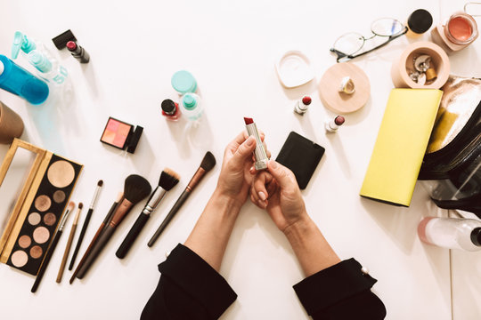 Close up make up artist holding red lipstick in hands with variety of cosmetic products on desk