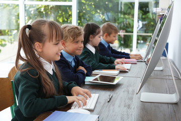 Little children in stylish school uniform at desks with computers - obrazy, fototapety, plakaty