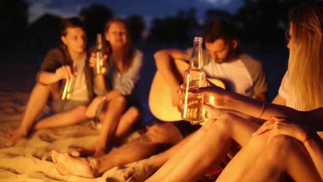 Beach party at sunset with bonfire. Friends sitting around the bonfire, drinking beer and singing to the guitar. Young men and women hold glass bottles with beverage singalong and cheering.