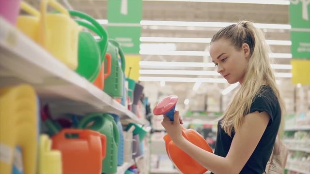 Blonde girl is examing a plastic watering can for pouring plants on a villa. She is standing in a hall of big store, preparing for summer season