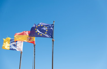 Canarias islands/ Spain-July 22, 2018: View from distance of tree flags of Spain, Europe Union and Canarias islands on blue sky and palms leaves background.