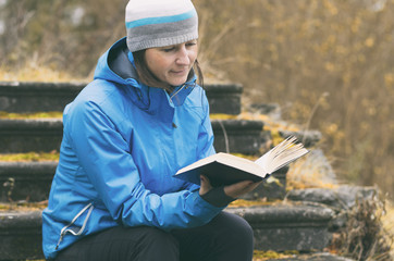 Obraz premium Autumn warmly dressed girl carefully reading a book on the background of yellow fallen leaves. Stylized film.