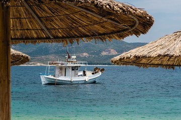 Summer lifestyle, vacation and travel concept. View from the beach trough the blured parasol made of cane, anchored sailing boat in focus and beautiful sea and sky in the background.