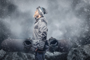 Brutal redhead snowboarder with a full beard in a winter hat and protective glasses dressed in a snowboarding coat posing with snowboard at a studio, looking away.