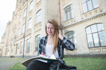 Portrait of a positive girl in a shirt sitting on a bench near a university building, reading a book and smiling. Attractive girl student is engaged in self-education at the university campus.