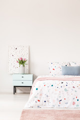 Flowers on pastel cabinet next to patterned bed with blue pillow in white bedroom interior. Real photo