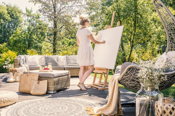 Young woman painting on a white canvas on a sunny terrace with garden furniture in the summer