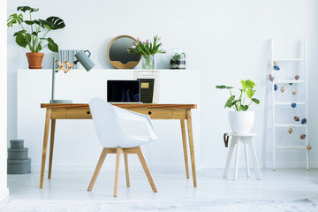Real photo of wooden home office desk with metal lamp and empty laptop standing in white living room interior with fresh plants and decor