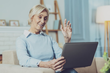 Video conversation. Pleased female person sitting on coach and using her laptop