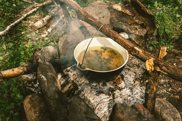 Herbal tea in cauldron is heats up on bonfire, surrounded by stones on background of ashes near...