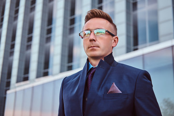 Close-up portrait of a confident handsome man in an elegant suit and glasses looking away while standing outdoors against a skyscraper background.