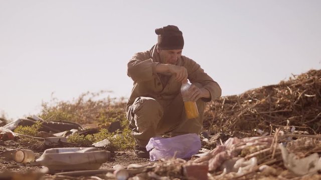 dirty homeless hungry man in a dump drinks the missing juice in the package with walking goes looking for food slow motion video. homeless dirty man roofless person looking for food in a dump. refugee