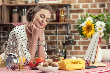 happy adult housewife with closed eyes leaning on table at kitchen