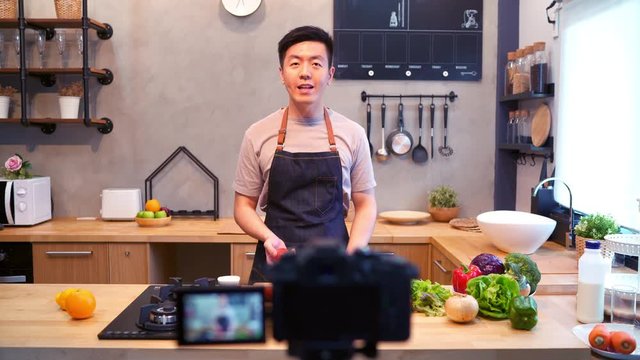 Young asian man in kitchen recording video on camera. Smiling asian man working on food blogger concept with fruits and vegetables in kitchen.