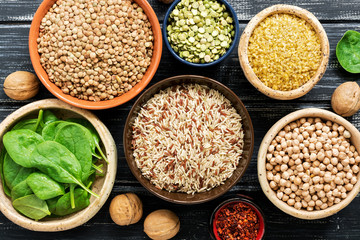 Chickpeas, lentils, peas, brown rice, bulgur, nuts and spinach on black rustic background. Various...