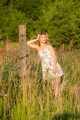 Fototapeta na wymiar Beautiful young woman standing in a field near a concrete pillar, green grass and flowers. Outdoors Enjoy nature. Healthy smiling girl standing in tall grass