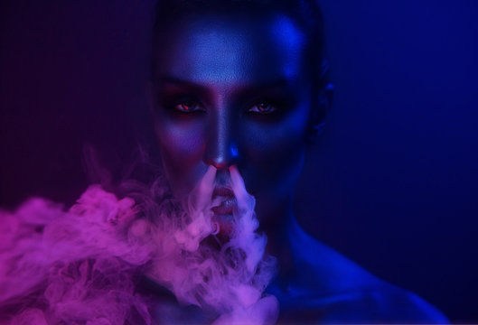 Nightlife. Beautiful Sexy Young Woman with glamorous mystical makeup vaping in Nightclub neon, exhaling smoke. Girl smoking