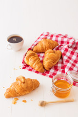French style breakfast, a white wooden table with cup of coffee, croissants and glass bowl of honey, few croissants placed on the red and white french checkered fabric.
