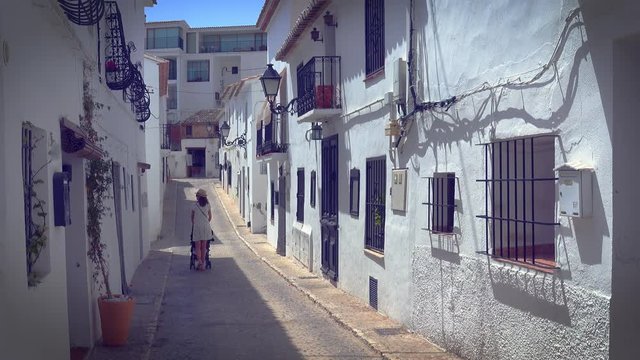 Mother strolling with newborn in in a narrow street with white houses in picturesque village of Altea, Alicante, Spain Vanishing point of view. Tourism and family travel concept