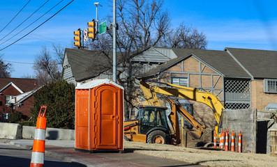Portable bathroom at construction site at intersectio of urban roads with backhoe in background and...