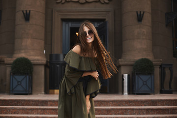 Naklejka premium Beautiful and fashionable brunette model girl with charming smile, in stylish dress with naked shoulders and in the trendy sunglasses posing outdoors in front of old building