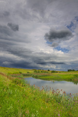 Sunny summer landscape with small river flowing between the green hills and fields.Beautiful dark clouds in dramatic overcast sky.