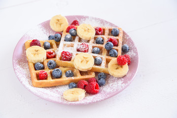 Belgian waffles with blueberries, raspberries, bananas and sugar powder on white background. Concept of tasty and healthy food.