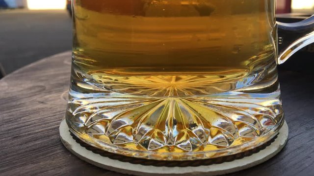 The bottom of a glass stein with golden colored beer, small carbonation bubbles rising from the center of the mug. Outside on a dark wood table.