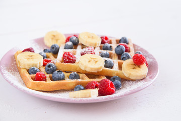 Belgian waffles with blueberries, raspberries, bananas and sugar powder on white background. Concept of tasty and healthy food.
