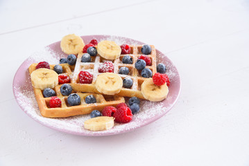 Belgian waffles with blueberries, raspberries, bananas and sugar powder on white background. Concept of tasty and healthy food.