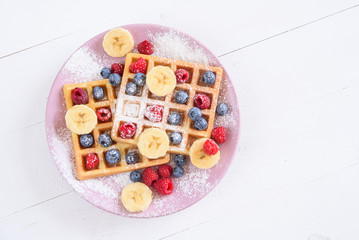 Belgian waffles with blueberries, raspberries, bananas and sugar powder on white background. Concept of tasty and healthy food. Top view.
