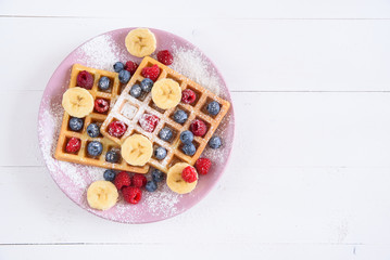 Belgian waffles with blueberries, raspberries, bananas and sugar powder on white background. Concept of tasty and healthy food. Top view.