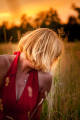 Portrait anonyme blonde Frau in rotem Kleid im Sonnenuntergang auf einer Wiese sitzend, makro...