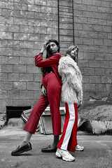 Growth portrait of two fashion young girl posing on the backyard. Stylish woman in a red suit and in white fur and sport red pants .on brick wall background.Black and white