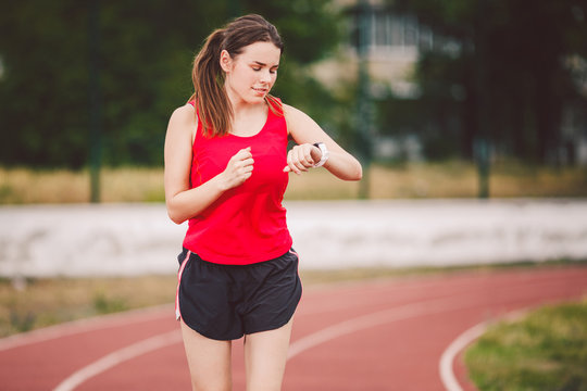 Beautiful young athlete Caucasian woman with big breast in red T-shirt and short  shorts jogging, running in the stadium. Hairstyle of a ponytail, uses a  sports watch on his arm Stock Photo
