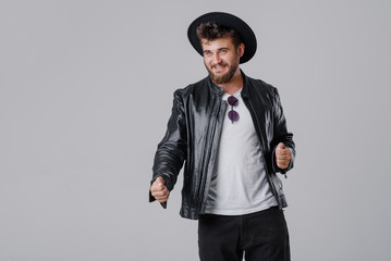 A young cheerful man with a beard in a stylish black leather jacket and hat on a gray background. Smiling at the good news