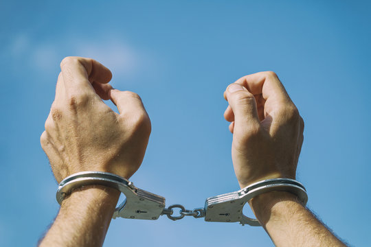 Hands of a man in handcuffs raised up and photographed against a blue sky background close-up