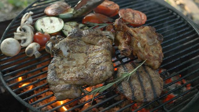 Assorted meat with grilled vegetables and rosemary sprigs, in the foreground hamburger cutlet, steak on the bone, ribs, grilled vegetables, zucchini, tomatoes, mushrooms, garlic head, chef turns the
