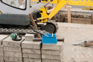 Strassenbau - Ein Gehörschutz und ein Hammer liegen auf Gehwegplatten aus Beton vor einem Minibagger