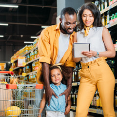 happy african american family with daughter looking at shopping list in supermarket